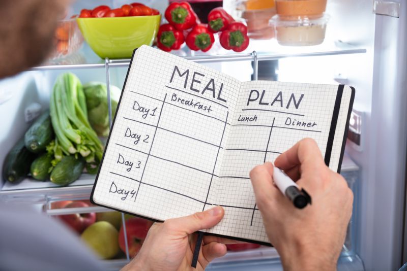 Wochenplan Essen erstellen Hände haöten geöffnetes Notizbuch in der Hand mit Menüplan, im Hintergrund geöffneter Kühlschrank